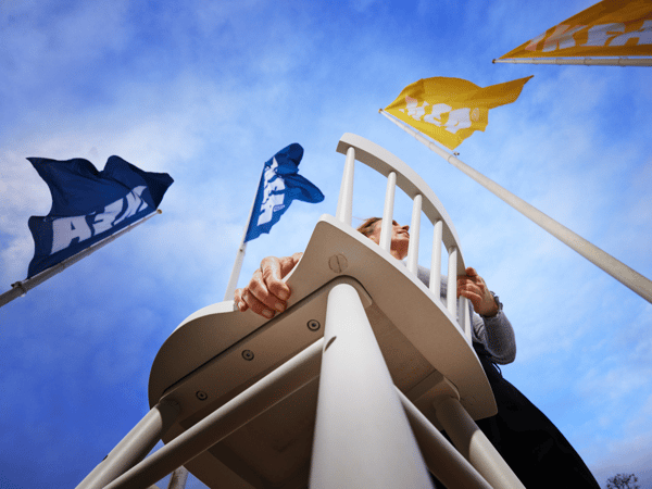 A person carries a white chair into an IKEA store, the IKEA flags are flying above them.