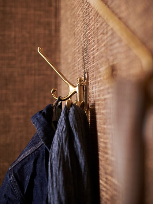 A brass-colour KÄMPIG three-arm hook attached to a light brown sea-grass wall. It holds a denim jacket and a dark blue scarf.