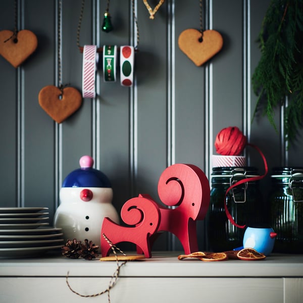 Två röda VINTERFINT-getprydnader står på ett sideboard bredvid en stapel tallrikar och andra festliga dekorationer.