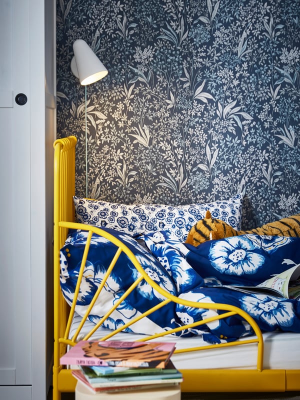 A white FUBBLA LED wall lamp shines on a child's yellow bed frame filled with blue-and-white bedding by a stack of books.