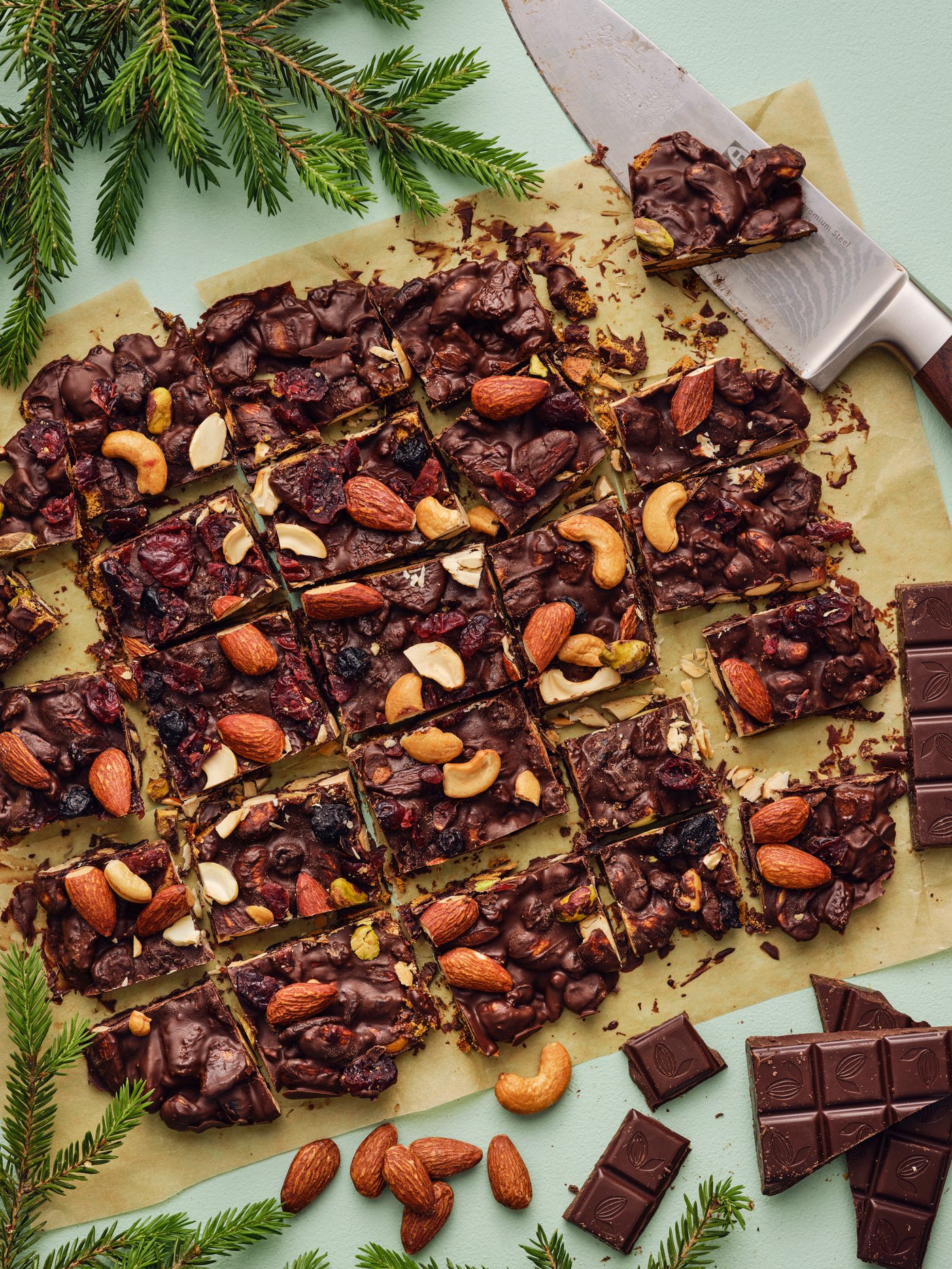 Vista dall'alto di una tavola con scorze di cioccolato con noci e frutti di bosco tagliate in porzioni. Accanto, del cioccolato, un coltello e rami di pino.