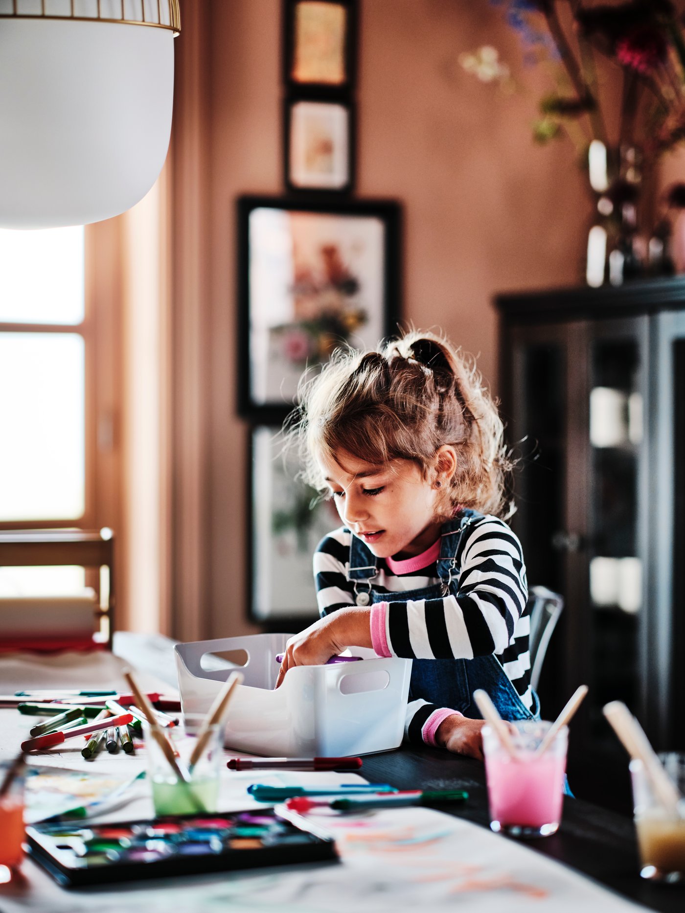 A child at a table covered in paper, pens and a mixed colour MÅLA watercolour box, taking a pen out of a grey VARIERA box.