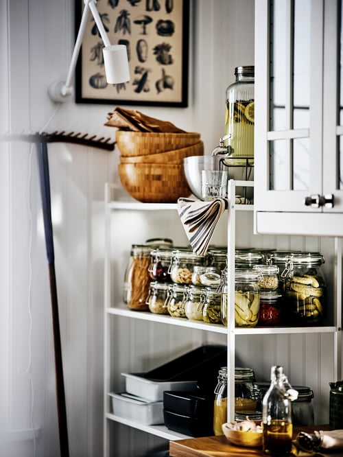 A white JONAXEL shelving unit holding jars of fruit, vegetables, kitchenware and wooden serving bowls against a white wall.