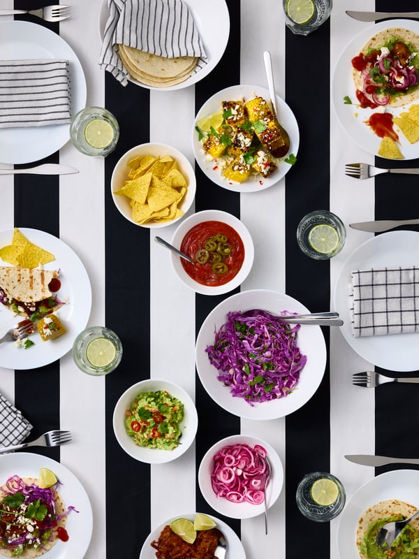 A table with a striped tablecloth has several white OFTAST bowls and deep plates with ingredients for a Mexican taco dinner.