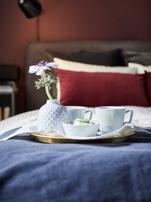 Bedroom featuring a round serving tray that contains a mug, a bowl, and a flower in a vase, all of which are placed on a bed.