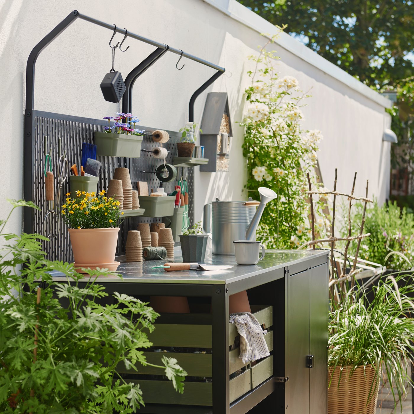 A black and stainless steel GRILLSKÄR kitchen island stands in a garden with green plants, pots and gardening tools on it.