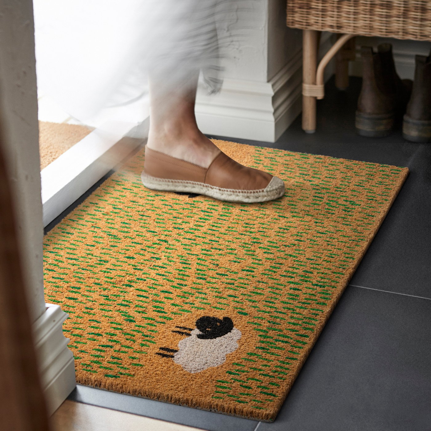 Woven doormat with a playful sheep design placed by a doorway, with a person stepping onto the mat in house slippers.