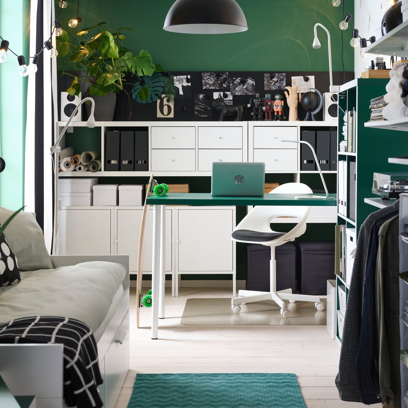 Una piccola stanza con un tavolo verde, scaffali bianchi, un letto divano, un guardaroba a giorno e una lampada a sospensione nera.
