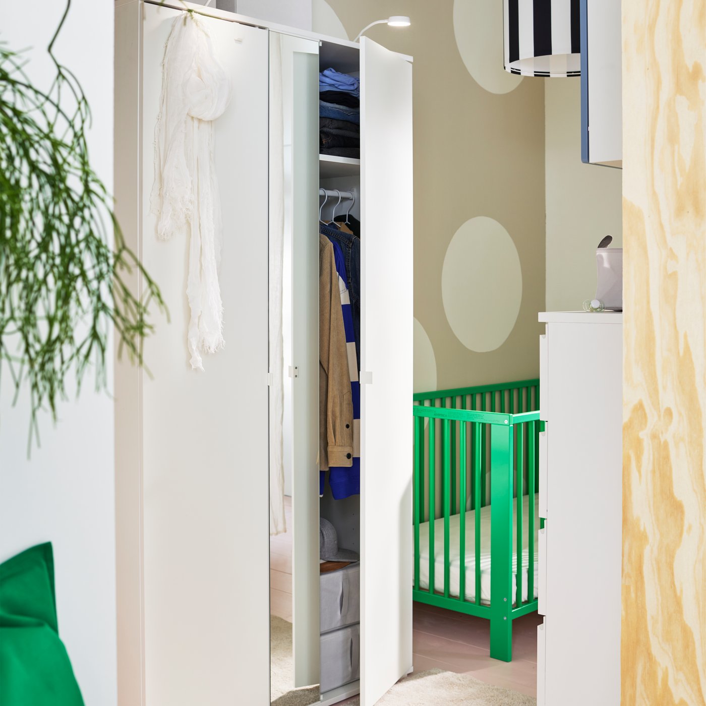 A white VIHALS wardrobe with 2 doors with one door slightly open showing hanging clothes and a green GULLIVER cot behind it.