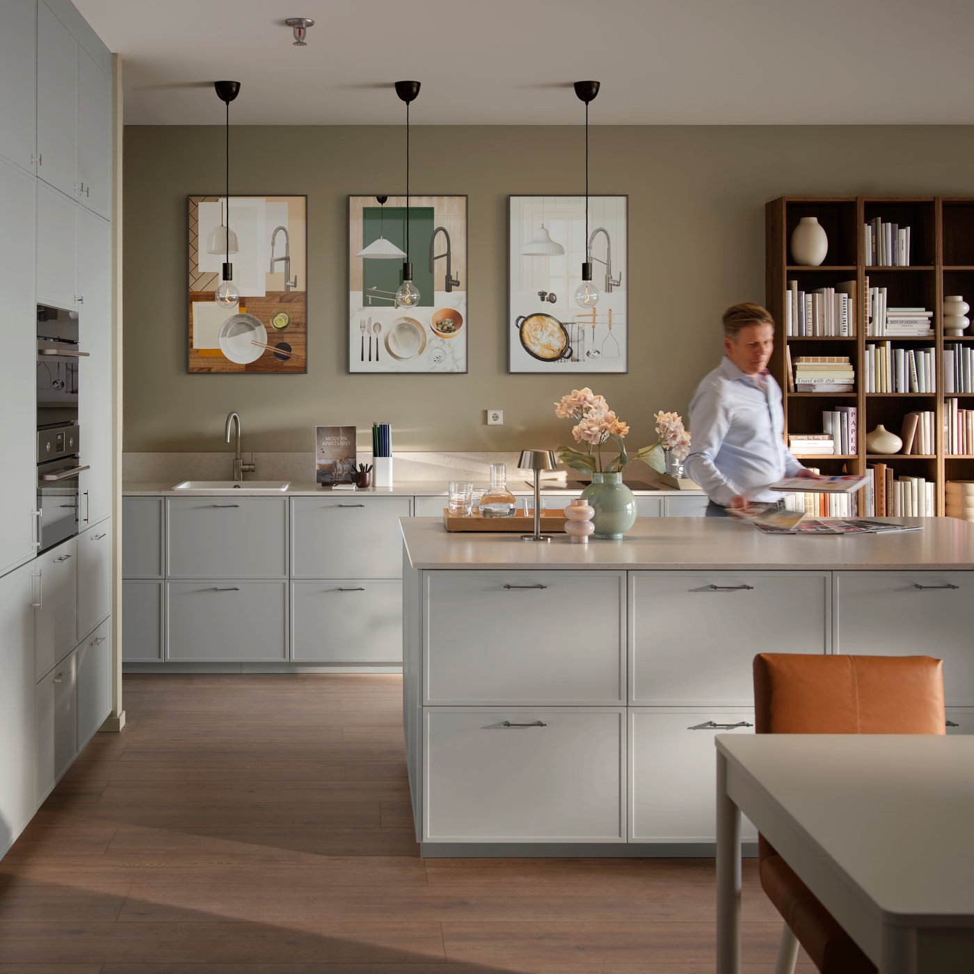 A model kitchen with light grey cabinets and built-in appliances. A man is leafing through brochures at a central island.