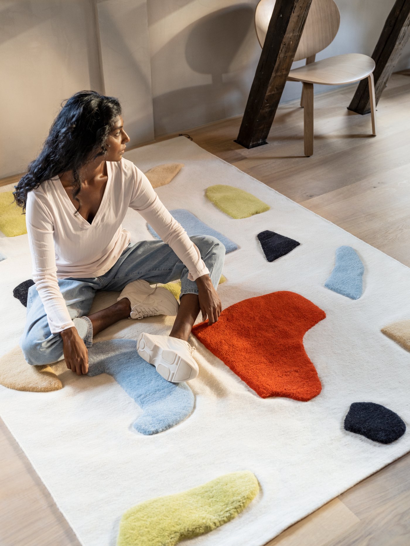 A young woman in jeans and a white top is sitting on a handwoven LOKALT wool rug with elevated colorful patches.