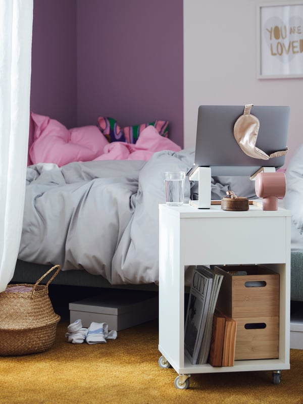 A laptop on a STENERIK laptop stand placed on a VIHALS bedside table next to a bed made with PILTANDVINGE bed linen.