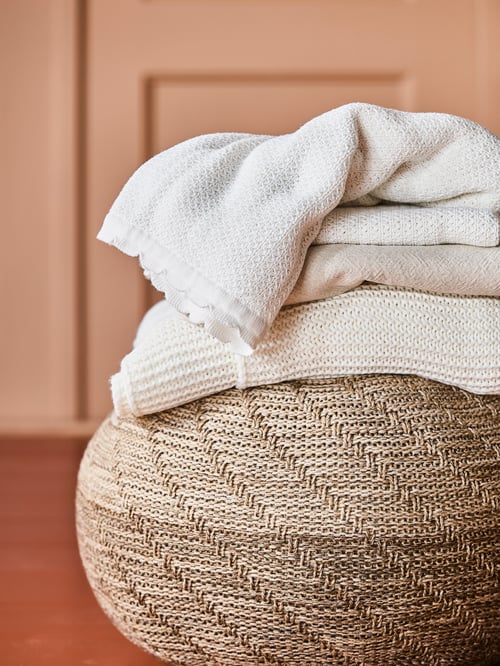 A pouffe has a stack of blankets on it, including a brioche-knitted, white cotton GULSPARV blanket with scalloped edges.