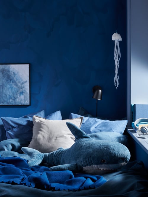 Section of a room with a rich, blue colour scheme, including a bed with a dark-blue POLARVIDE throw and a BLÅHAJ soft toy.