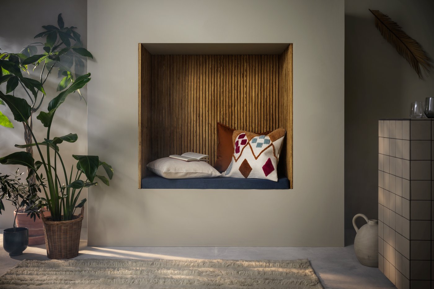 Una racó de lectura vora una paret decorat amb coixins al costat de testos amb plantes frondoses a una banda i un gerro a l'altra.