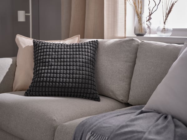 I en lys beige sofa ligger en pude med et GURLI betræk, en pude med et sort SVARTPOPPEL pudebetræk og et HOLMVI tæppe slænget henover sæddet, bag ved sofaen anes et HÄLLEBRÄCKA gardin i en stue.