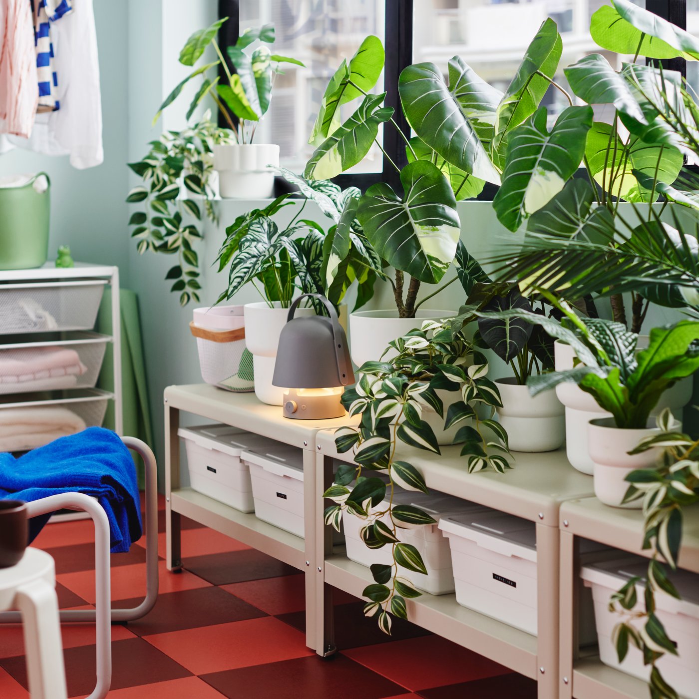 Lots of artificial potted plants like FEJKA tradescan zebrina are on shelving units below the windows of an enclosed balcony.