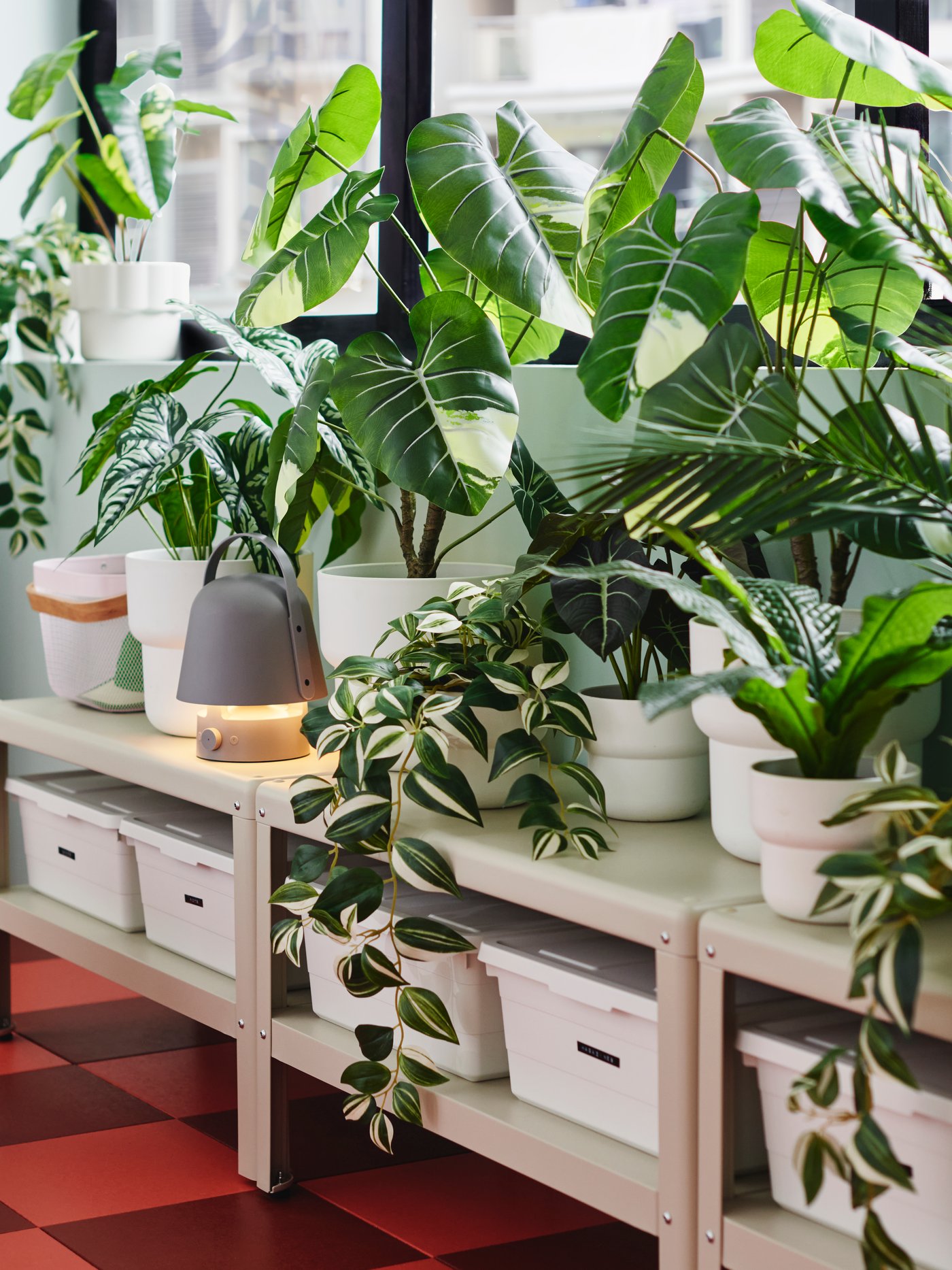 Lots of artificial potted plants like FEJKA tradescan zebrina are on shelving units below the windows of an enclosed balcony.