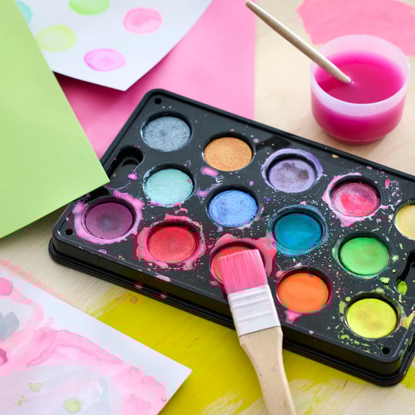 Close-up of MÅLA watercolour box, a brush, a cup of water and different coloured paper underneath.