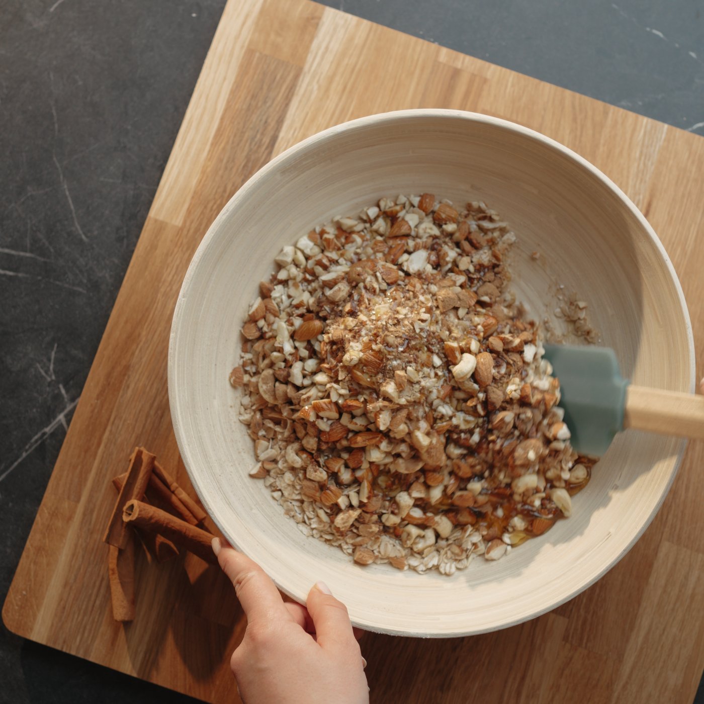 La mano di una donna mescola un composto di noci e spezie in una ciotola bianca su un tagliere di legno. Accanto, su un piano di lavoro scuro, sono appoggiati dei bastoncini di cannella.