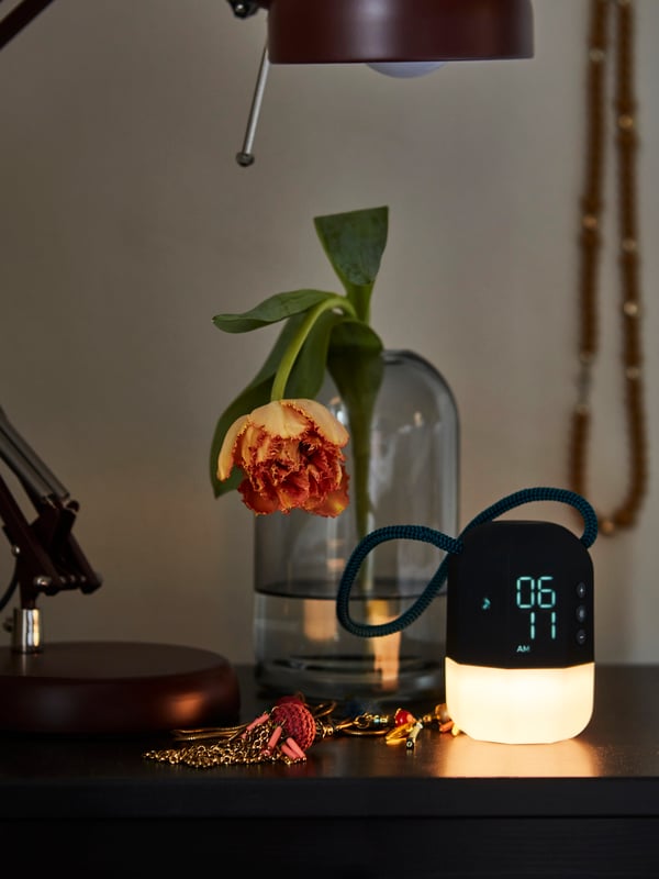The top of a bedside table with a table lamp, some jewelry, a vase with a flower and a FNURRA alarm clock/wake-up light.