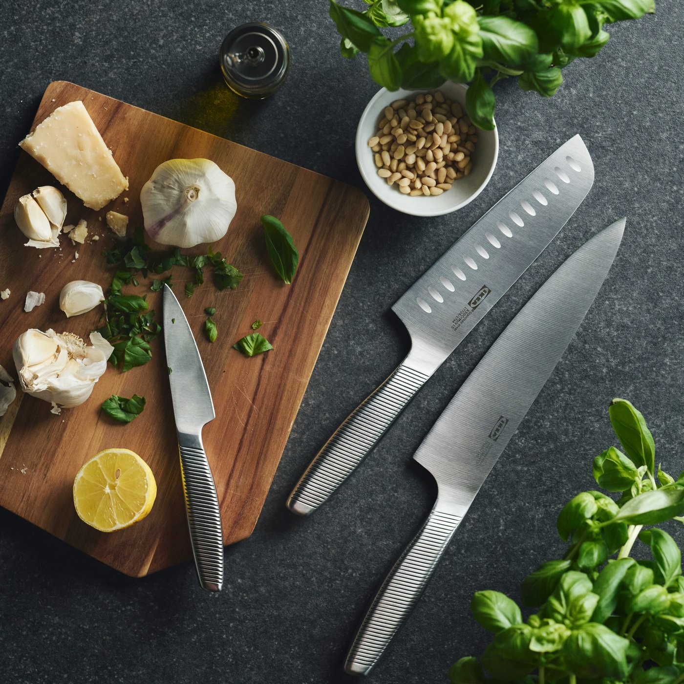 Three IKEA 365+ knives in various sizes on a kitchen worktop surrounded by basil, parmesan cheese, garlic and pine nuts.