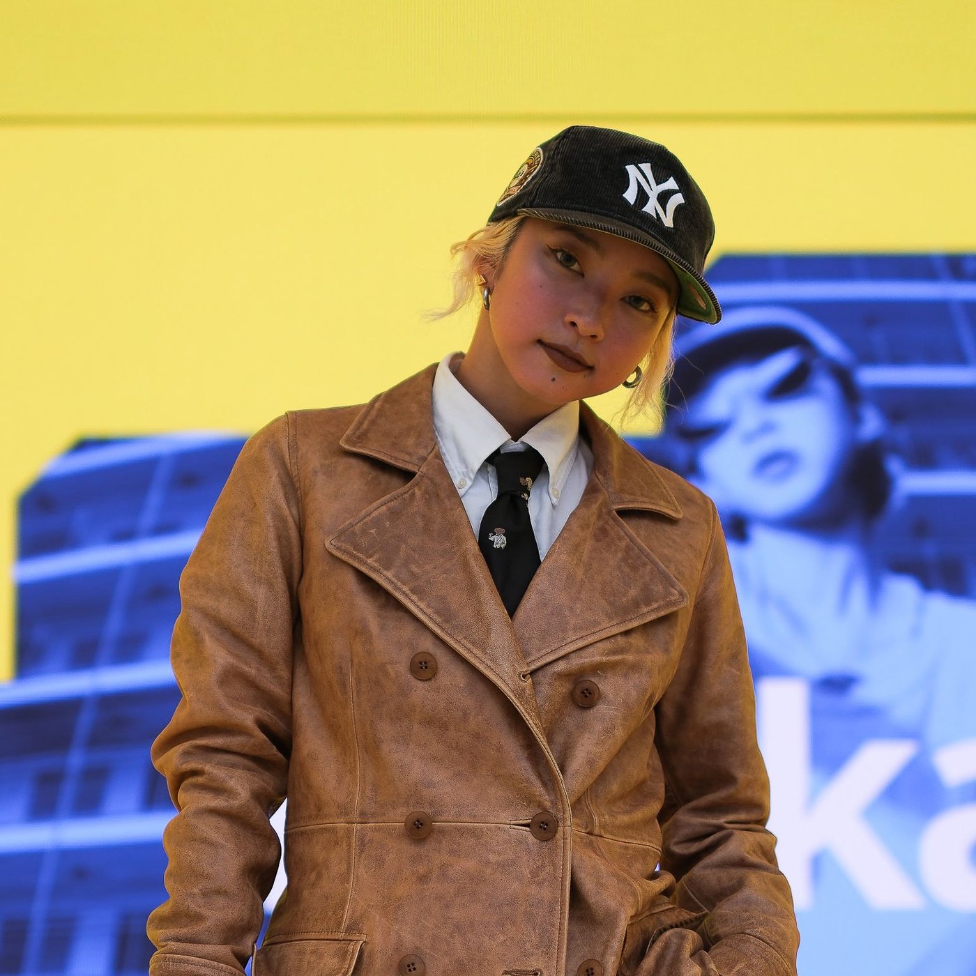 Während der Mailänder Designwoche 2024 steht Kyoka am IKEA Standort auf einer Bühne. Sie trägt einen Ledermantel, einen Hut, Krawatte und ein weisses Hemd. Im Hintergrund ist eine digitale Anzeige in Blau und Gelb zu sehen.