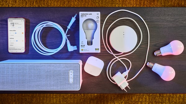A white SYMFONISK WiFi bookshelf speaker, a white TRÅDFRI gateway, a white remote control and a cable.