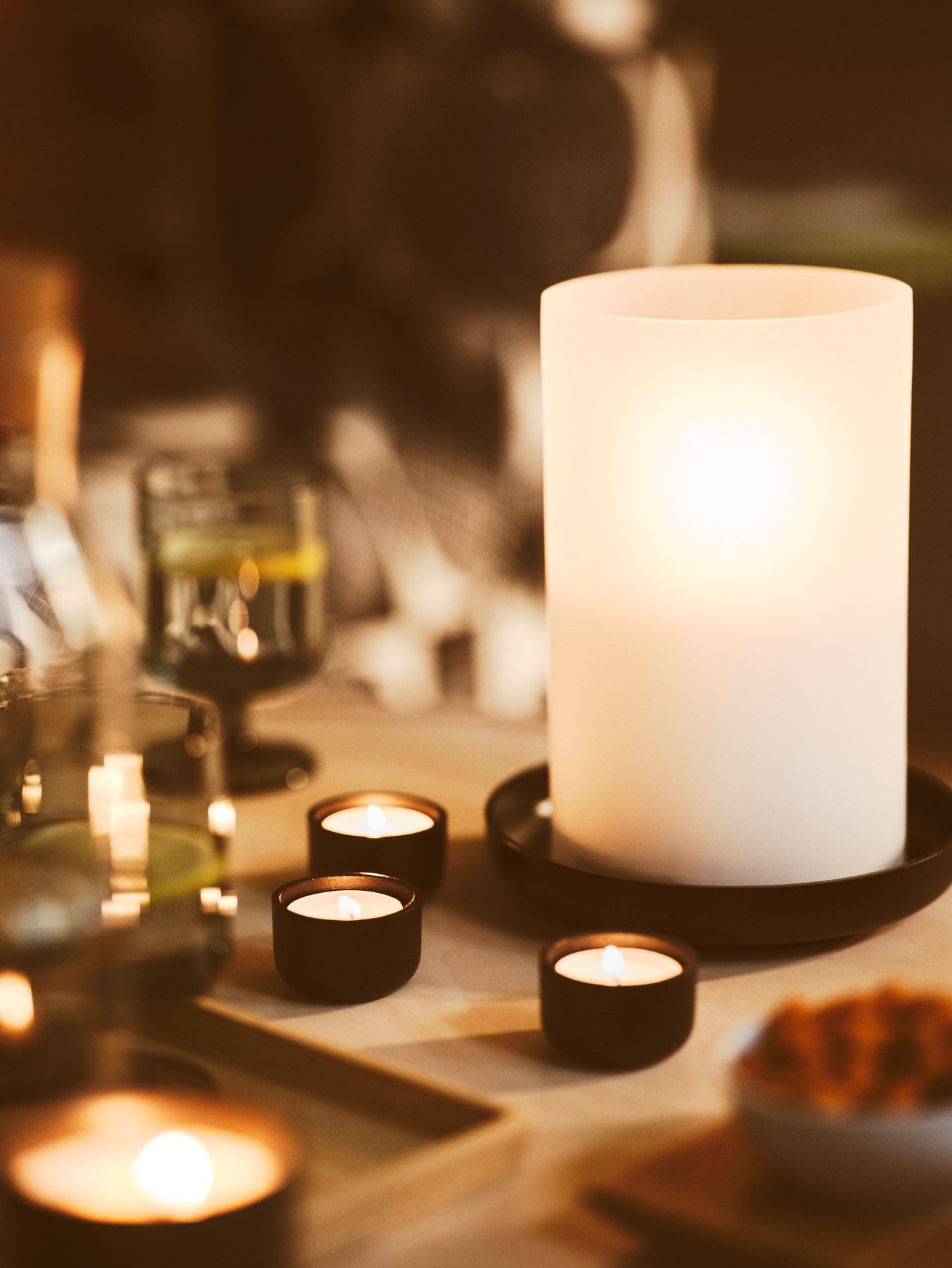 Unos portavelas pequeños HEDERVÄRD negros con velas encendidas están situados junto a un farol HEDERVÄRD encendido, al lado de unas copas y una bandeja.