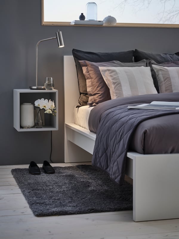 Una cama MALM blanca con ropa de cama en tonos grises en una habitación minimalista. Al costado de la cama una bajada de cama gris LINDKNUD.