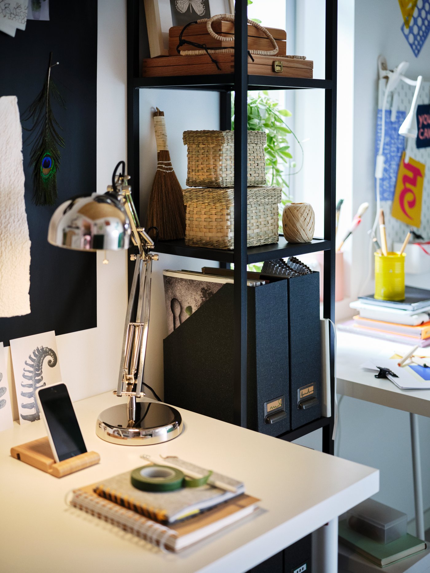 Next to a desk with a white LAGKAPTEN tabletop lit by a FORSÅ work lamp, stands an anthracite ENHET five-shelf high frame.