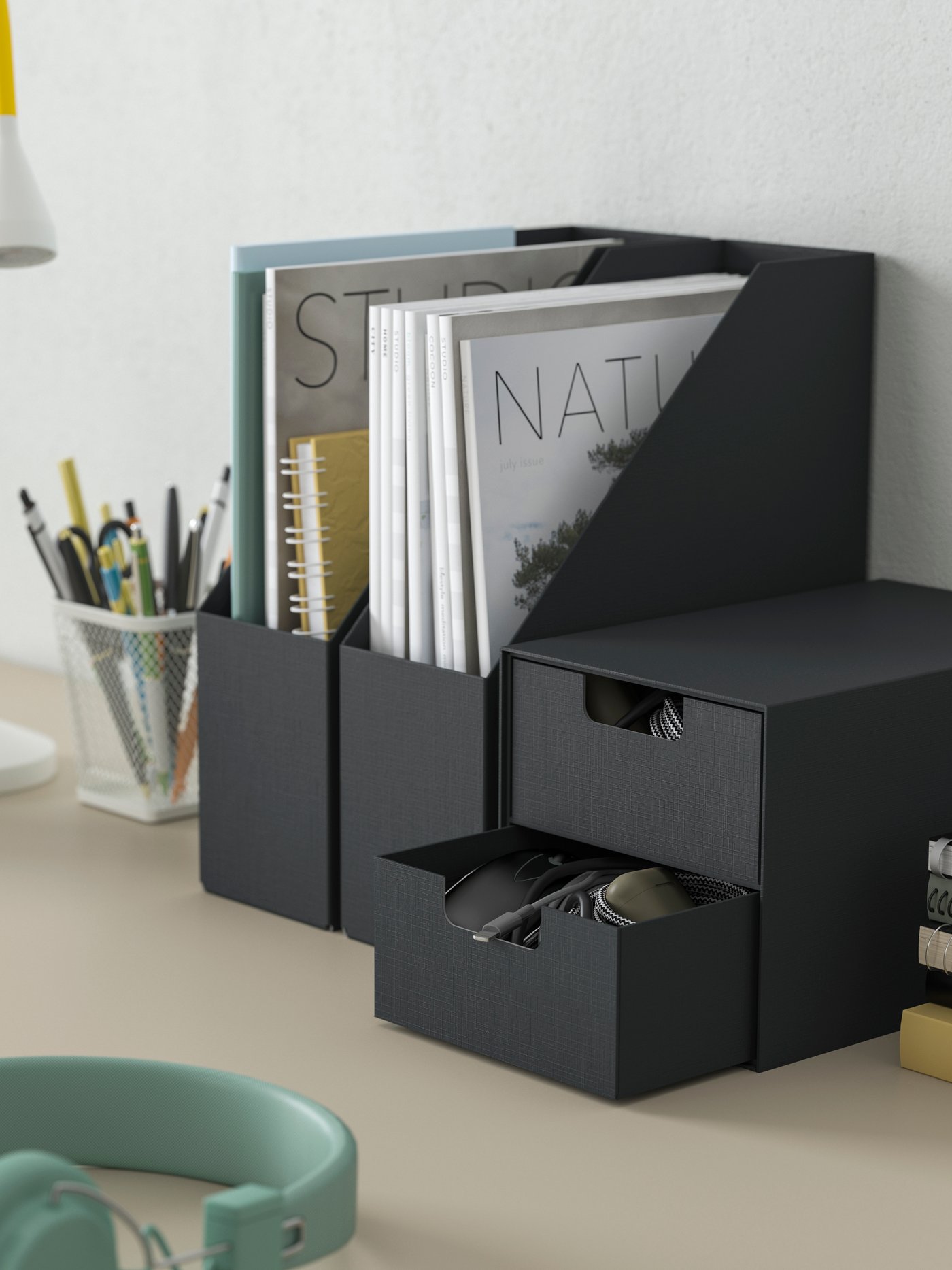 Beige skrivbordsskiva där två stycken svarta FJÄDERHARV tidskriftshållare och en minibyrå med två lådor står placerade mot den vita väggen. På skrivbordet syns även en vit och gul skrivbordslampa, ett vitt pennställ och ett par mintgröna hörlurar.