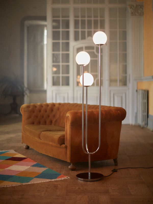 Soggiorno con pavimento in legno, termosifone, tappeto a tessitura piatta VINDERÖD, divano marrone e lampada da terra SIMRISHAMN.