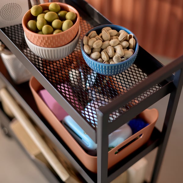A close-up of a black NISSAFORS trolley that has items stored on the mesh shelves, including bowls of nuts and olives.