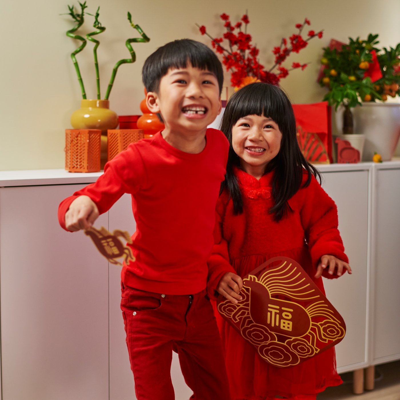 Two children dressed in red, in front of a white EKET wall-mounted shelving unit, holding a FÖSSTA
red window decoration shaped like a horse