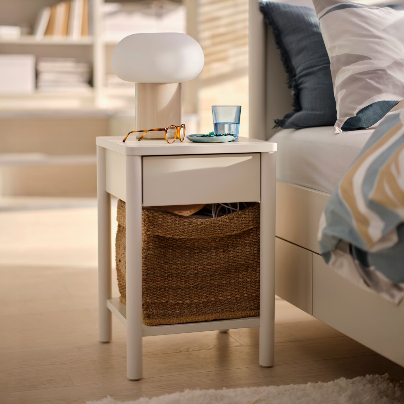 White bedside table next to white bed, with a basket on the lower shelf, a table lamp and glass of water on top.