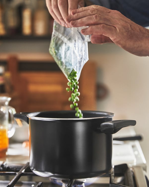 Pentola HEMLAGAD nera con rivestimento antiaderente posta su un fornello a gas mentre una persona sta rovesciando un alimento al suo interno.