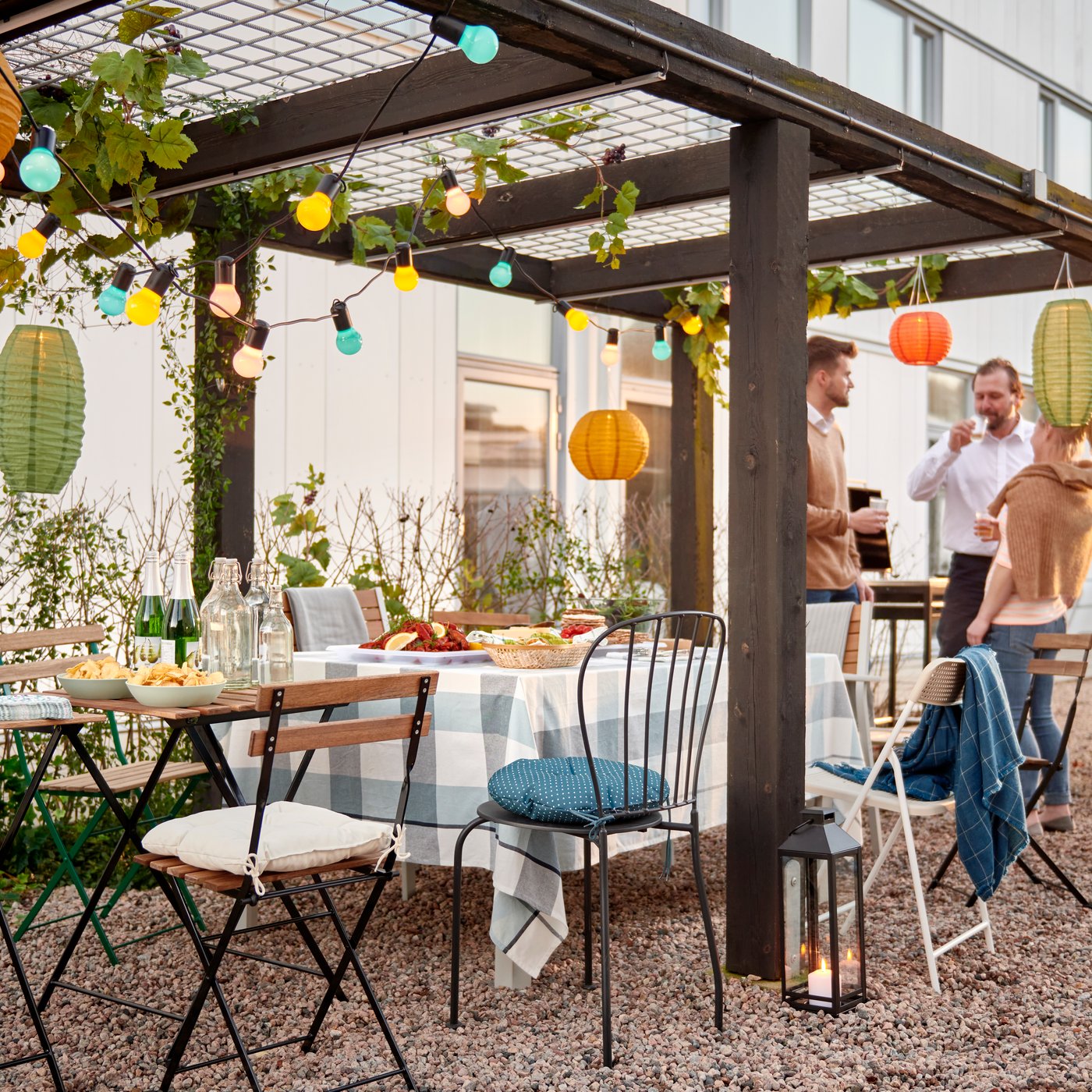 A group of people standing by a row of chairs and small tables set for a party under a decorated pergola in a courtyard.