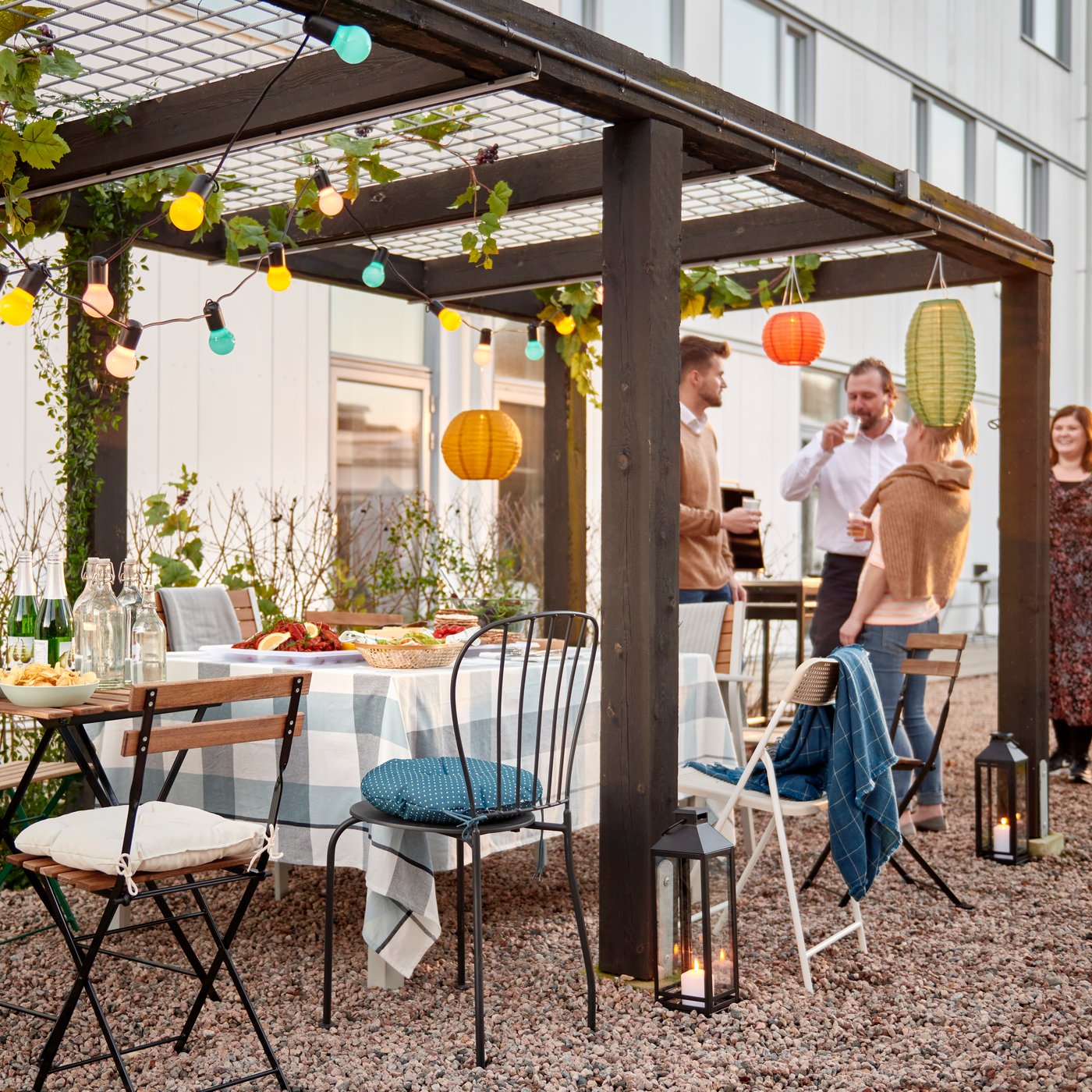 Un gruppo di persone in piedi, vicino a una fila di sedie e a dei piccoli tavoli TÄRNÖ apparecchiati con una tovaglia RUTIG per una festa sotto una pergolato decorato in un cortile.