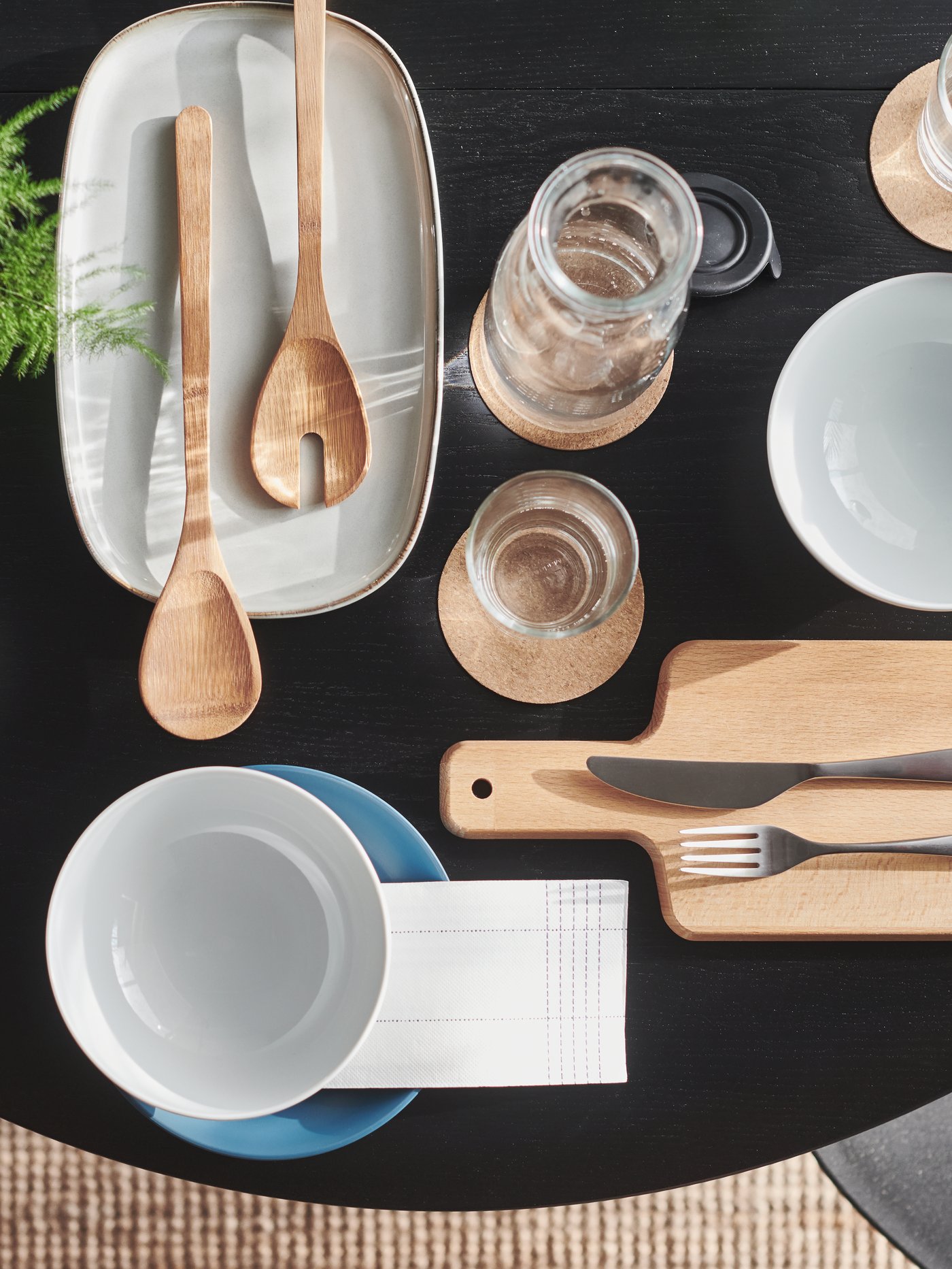 Vista superior de un plato gris con dos utensilios para servir ensalada, una jarra con tapa, un cuenco blanco y una tabla de cortar con cubiertos.
