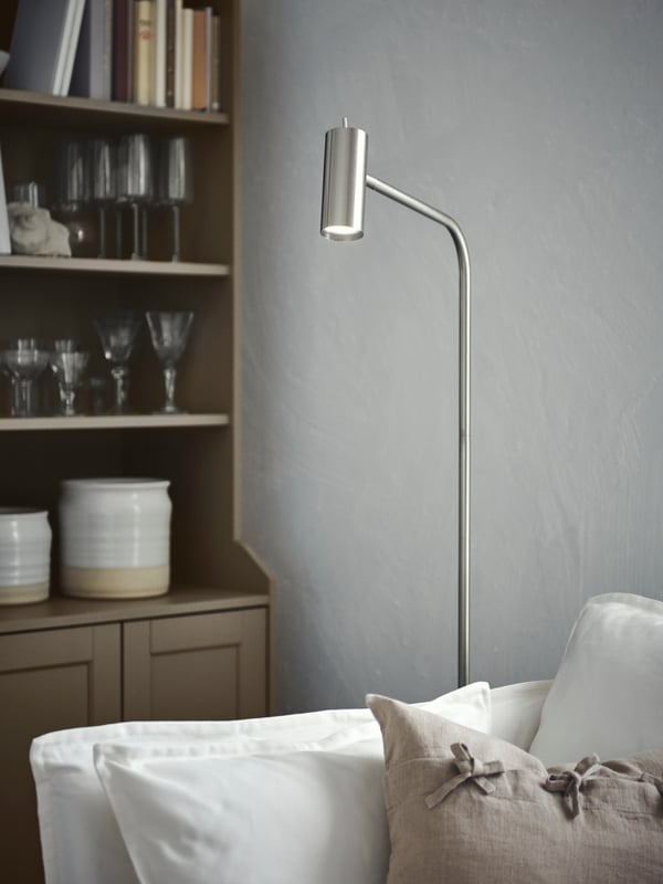 A corner with a lit nickel-plated VIRRMO floor lamp standing between a white BACKSÄLEN armchair and a beige HAUGA cabinet.