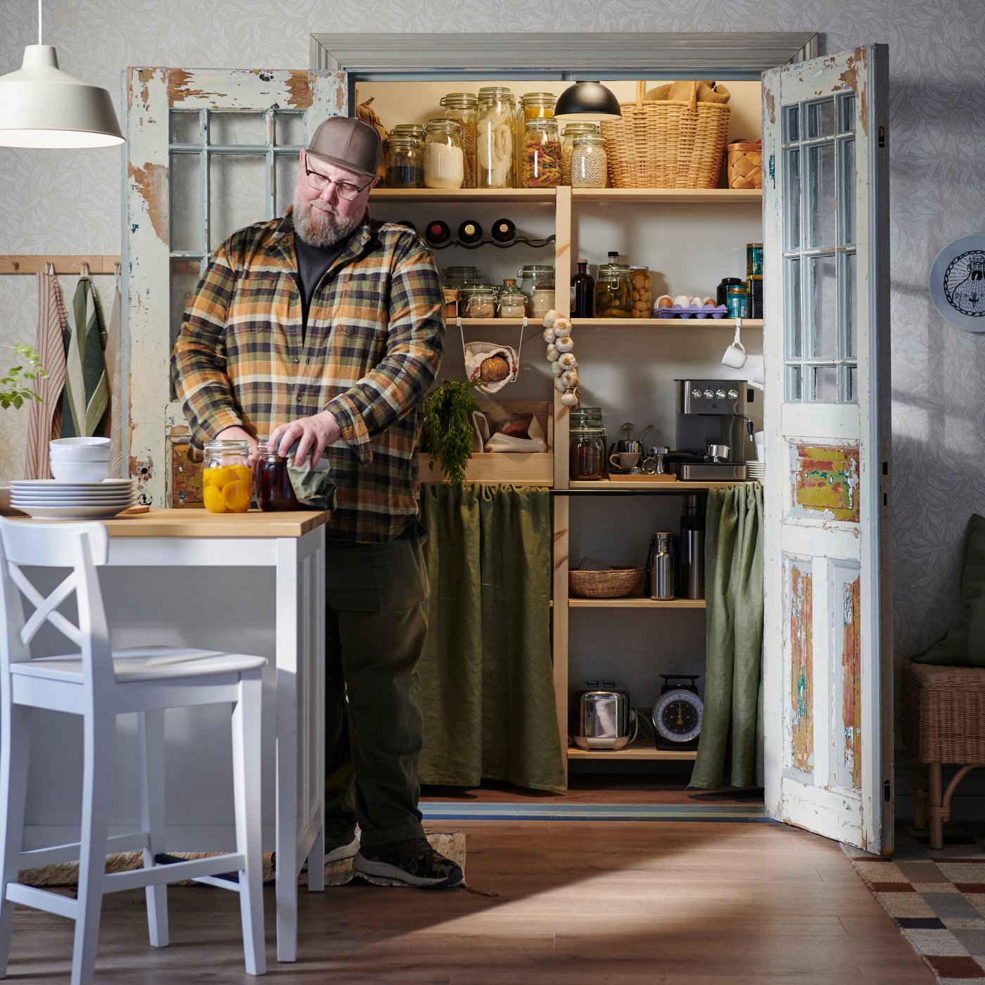 A large man in a plaid shirt and baseball cap stands in front of his walk-in pantry considering several jars of pickles.