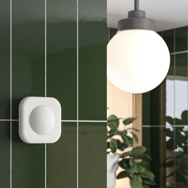 A VALLHORN wireless motion sensor is placed on a bathroom wall close to a FRIHULT ceiling lamp in stainless steel colour.