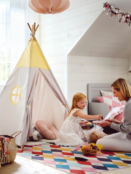 A girl sitting on the floor by a HÖVLIG play tent holding a plate of toy cakes giving one to her mum sitting opposite her.