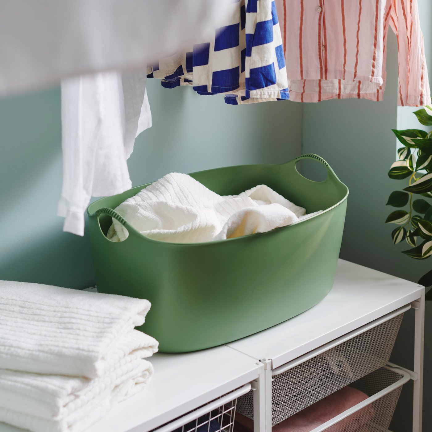 Na beli površini za zlaganje perila, je zelena TORKIS mehka košara za perilo s prostornino 35 litrov, primerna za notranjo in zunanjo uporabo, v njej pa so brisače.