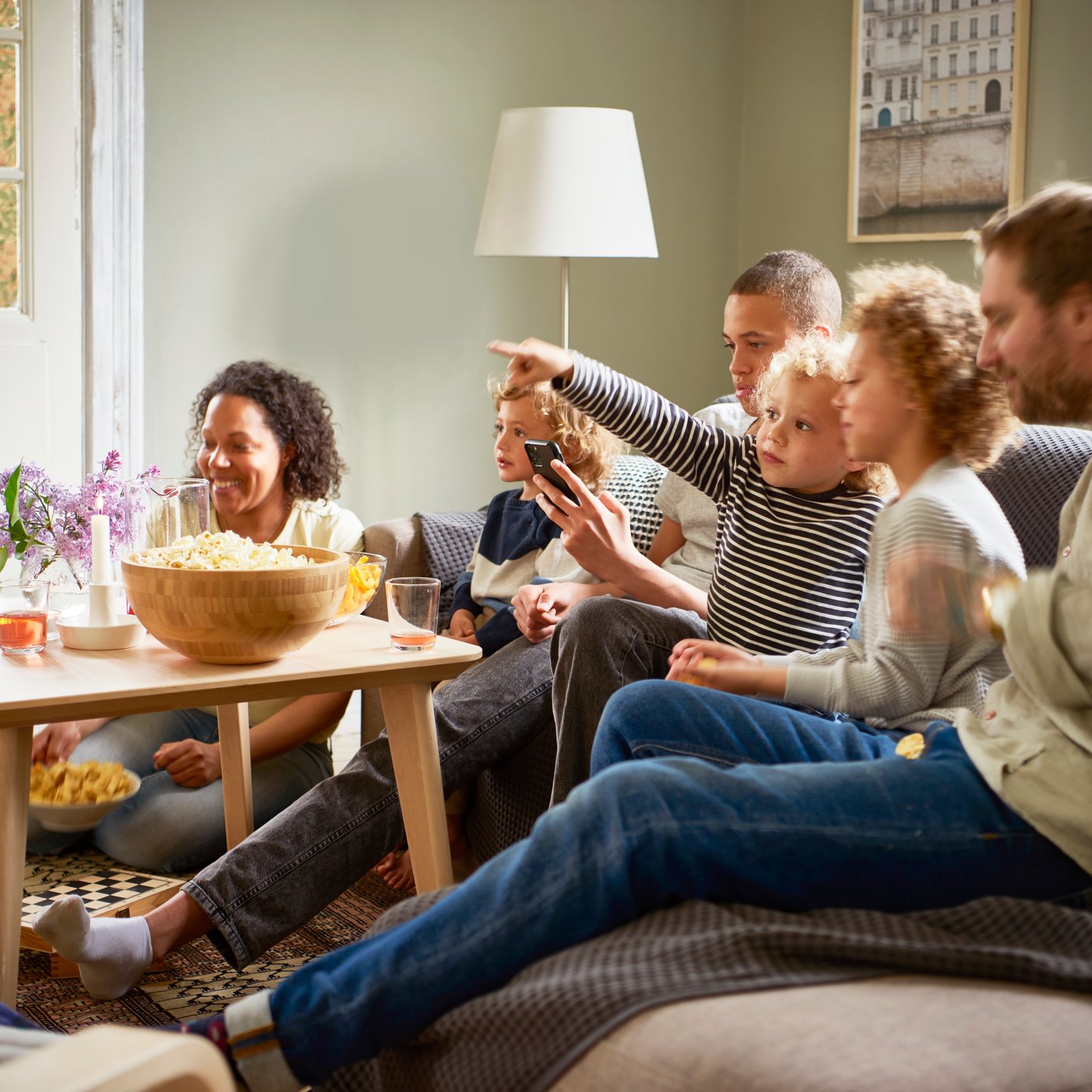 A family sitting together on a sofa, happily chatting with one another, with a bamboo bowl filled with popcorn on the coffee table.