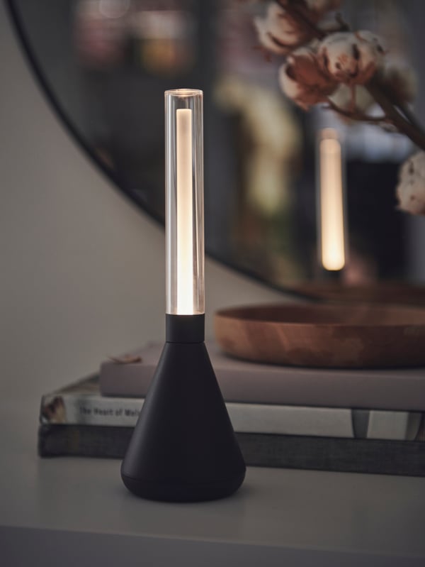 A dim room with a lit black BJÖRKSPIREA tube-shaped LED light decoration, its light reflected in a nearby LINDBYN mirror.