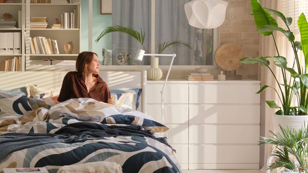 A person is sitting in a bed made with MYGGLASVINGE bed linen in a bright bedroom with chest of drawers, plants and more.