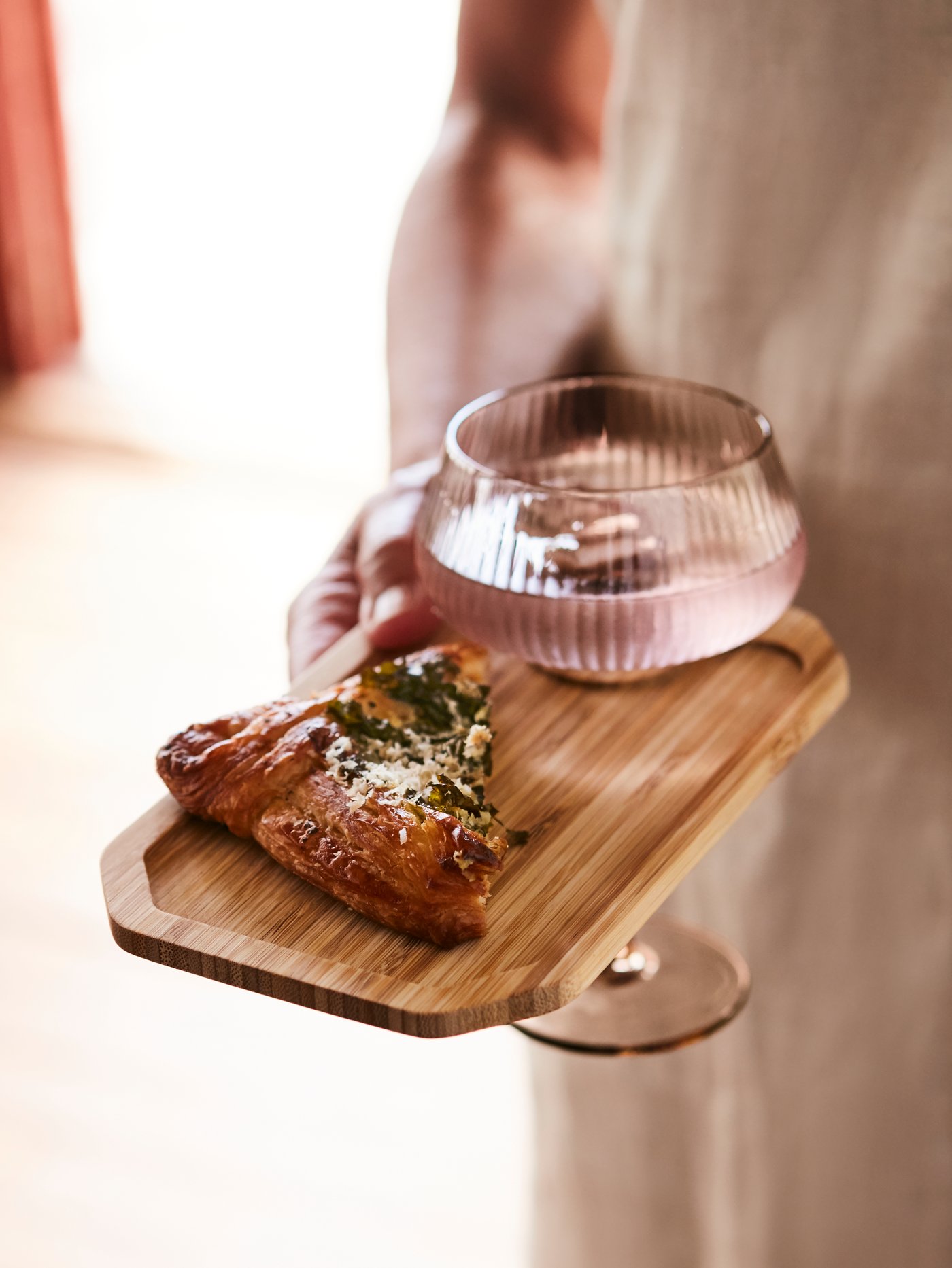An ANLEDNING mingle plate made in bamboo holding a glass and a savoury item, held by a woman in a beige dress.