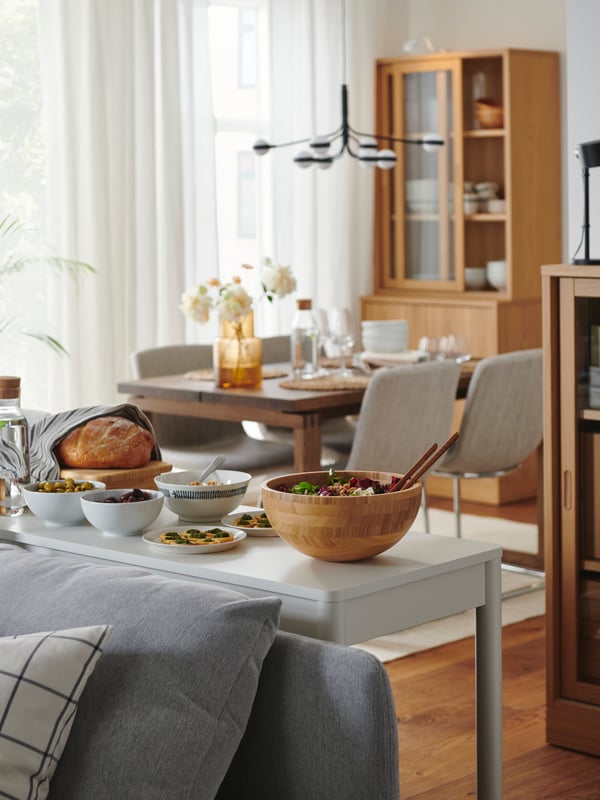 A modern living room with a dining area. A TONSTAD desk is placed behind a sofa and is used as a prep area for food.